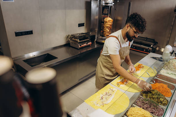Male chef in apron and gloves preparing shawarma in a commercial kitchen, adding fresh ingredients to flatbreads for middle eastern street food