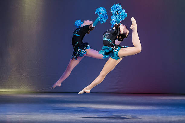 Two young cheerleaders perform a synchronized jump with high leg extensions, holding blue pom-poms mid-air. Their dynamic pose, strength, and flexibility are highlighted against a colorful studio background.
