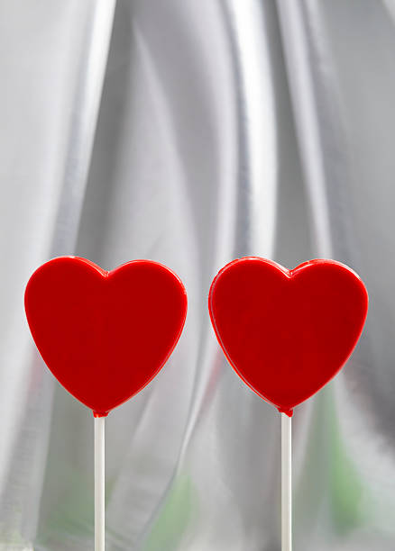 Minimalist composition featuring two red heart-shaped lollipops on a soft fabric background. The clean design and balanced symmetry make this image ideal for graphic design projects, website headers, social media graphics, print layouts, and marketing designs requiring a bold love symbol with visual simplicity.