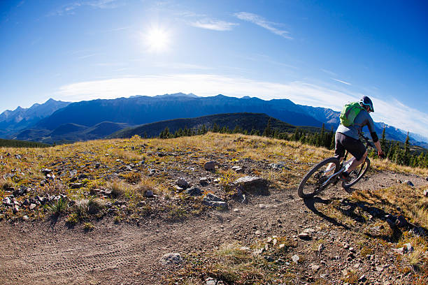 A man rides his full-suspension, electric-powered mountain bike down a singletrack trail in the Rocky Mountains of Alberta, Canada, on a sunny day. He wears cycling clothing, a backpack, and a bicycle helmet.