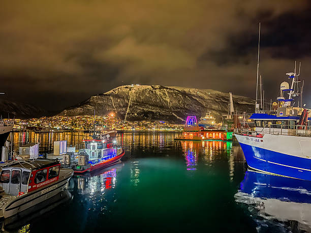 Tromso, Norway - December 21, 2025: Winter polar night. Looking east to the mainland with boats docked downtown. Black sky and many lights on houses and the fishing boats