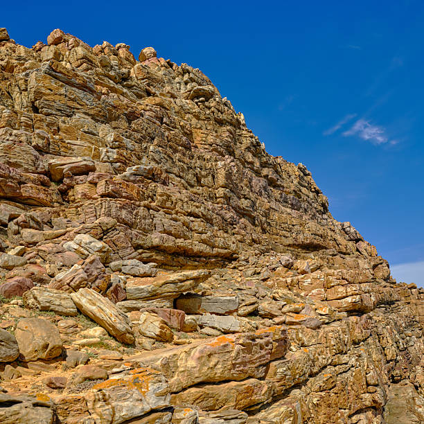 A stunning Western Cape landscape featuring a towering, layered rock cliff and scattered boulders. Sunlit textures reveal rich earth tones and geological strata against a vivid blue sky, illustrating rugged nature and dramatic coastal-hinterland terrain perfect for travel, nature, and geology themes.