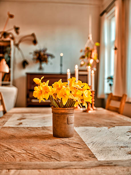 A charming still life featuring a bouquet of Narcissus pseudonarcissus in a rustic metal vase positioned at the center of a wooden dining table. Warm candlelight and a softly blurred background create a homely, inviting atmosphere typical of a Danish interior. Ideal for home decor, spring, and warmth themes.