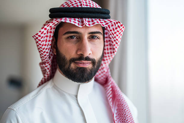 Focused young Arabic businessman wearing traditional keffiyeh and thawb, standing confidently by a window in an office setting, reflecting a professional and optimistic demeanor.