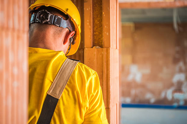 A construction worker wearing a yellow helmet and shirt is focused on tasks within a partially constructed building. The environment shows unfinished walls and tools, indicating ongoing work.