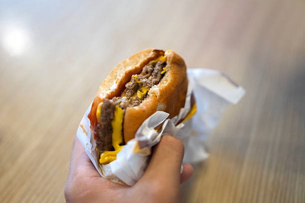 Close-up of a hand holding a partially eaten cheeseburger with a beef patty, melted cheese, and mustard sauce wrapped in paper.