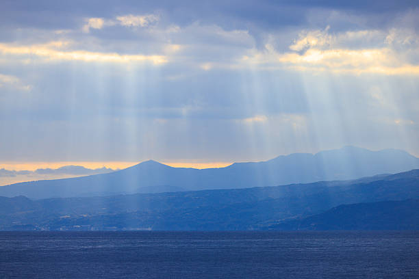 Seascape view of soft sun rays piercing dense clouds above calm gray-blue sea, captured in natural daylight from shore with horizon line and atmospheric mood in cloudy weather.
