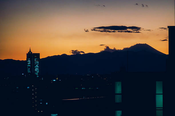 A calm dusk view of Mount Fuji appearing as a dark silhouette beyond the city skyline of Tokyo.The fading orange and blue sky creates a tranquil atmosphere, symbolizing the contrast between urban life and nature in Japan.