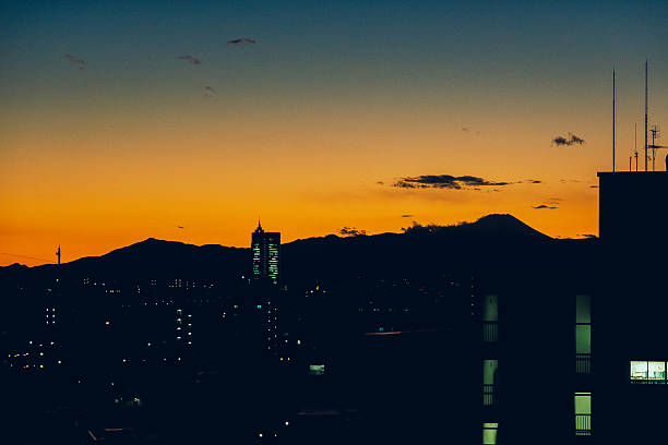 A calm dusk view of Mount Fuji appearing as a dark silhouette beyond the city skyline of Tokyo.The fading orange and blue sky creates a tranquil atmosphere, symbolizing the contrast between urban life and nature in Japan.