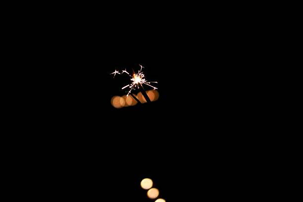 Minimal composition of a sparkler bursting with bright sparks in the dark night. Small light source surrounded by deep black background, creating strong copy space for celebration, concept, and abstract design use.