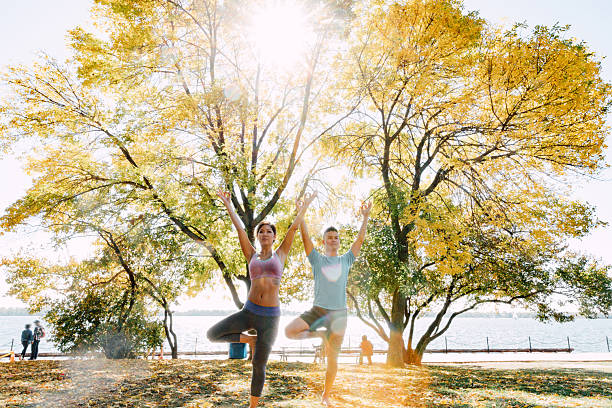 Athletic man and woman practicing partner yoga and acroyoga poses on a wooden deck near the water during daylight. The couple performs balance, stretching, and strength exercises together, including assisted poses and lifts, emphasizing trust, coordination, and physical connection. Outdoor wellness activity combining fitness, mindfulness, and teamwork in a calm urban waterfront environment.