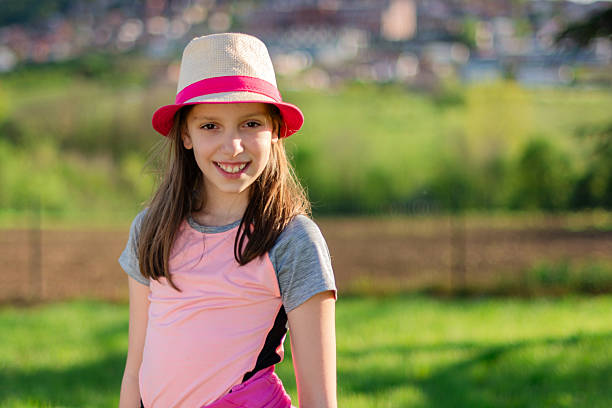 Smiling girl wearing a hat and pink shirt outdoors, enjoying a bright sunny day. Childhood happiness in nature