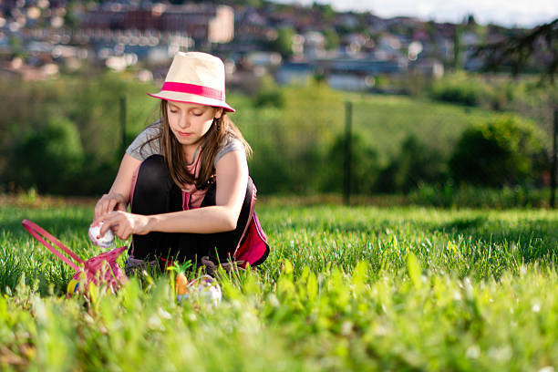 Girl crouching on green grass, wearing a hat, finding Easter egg in her pink basket during a sunny spring day outdoors