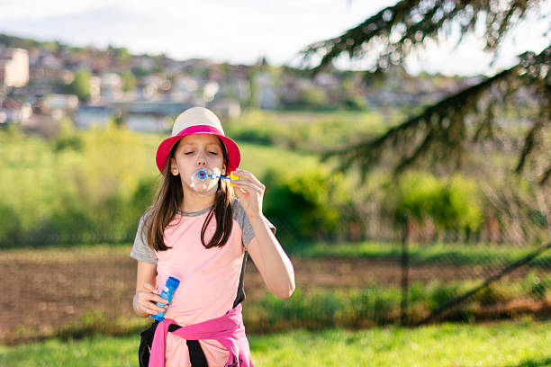 Young girl wearing a hat blowing colorful soap bubbles outdoors in a green park enjoying childhood and summer fun