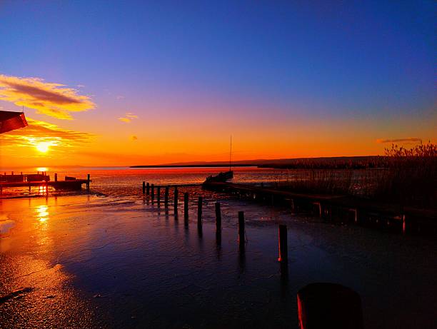 Sun setting behind boat dock at Neusiedler See Seegelhafen West, silhouetting small boat, wooden posts and reed beds with golden reflections on shallow icy water amid purple-orange sky and clouds.