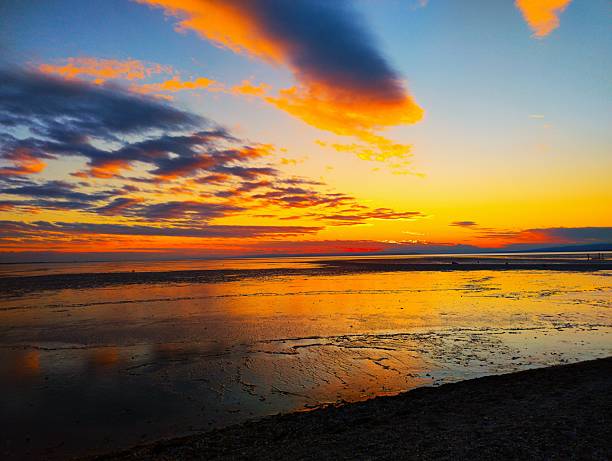 Wide shot capturing a calm ocean horizon during sunset with warm orange and blue tones in the sky, a crescent moon visible above, and a quiet beach in silhouette creating a peaceful coastal atmosphere.