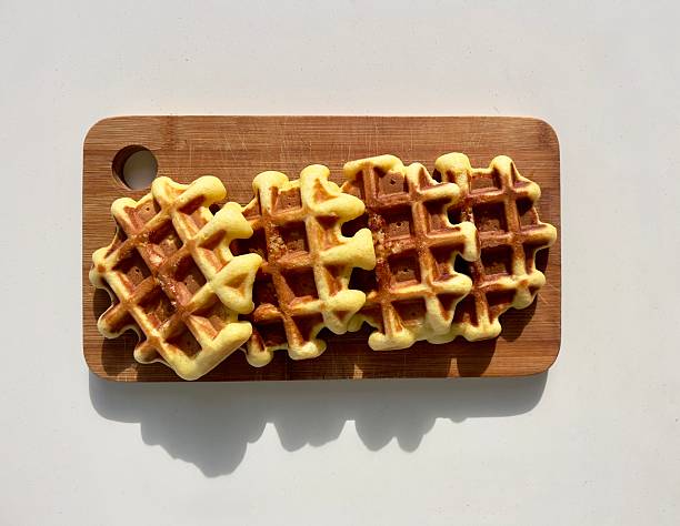 Freshly baked golden waffle with crisp edges and soft texture on wooden board, showing detailed waffle grid and melted sugar. Warm lighting highlights the crunchy surface and appetizing homemade breakfast feel