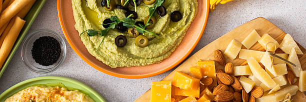 Broccoli hummus is artistically displayed in a bowl, garnished with olives. Accompanied by a charcuterie board featuring cheese, nuts, and breadsticks for a delightful snack experience, banner