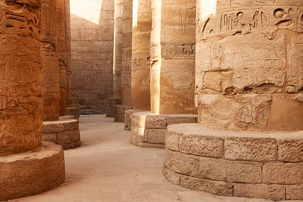 A sunlit walkway between massive sandstone pillars carved with intricate hieroglyphs and bas-relief figures. The worn, rounded column bases and sandy floor show centuries of erosion, while warm light and deep shadows emphasize the texture and scale of the stonework. The scene conveys a quiet, archaeological atmosphere inside a monumental ancient temple.