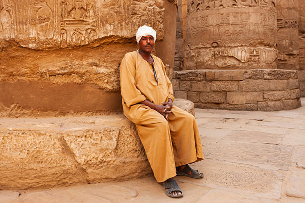 A man in a loose, ochre-colored robe and white turban sits against a weathered stone wall carved with hieroglyphs, hands clasped and feet in sandals. Warm sandstone tones and massive carved columns create a quiet, timeless scene at an ancient temple.