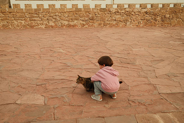 Young child squatting, gently stroking a tabby cat on a textured stone floor with an old stone wall in the background
