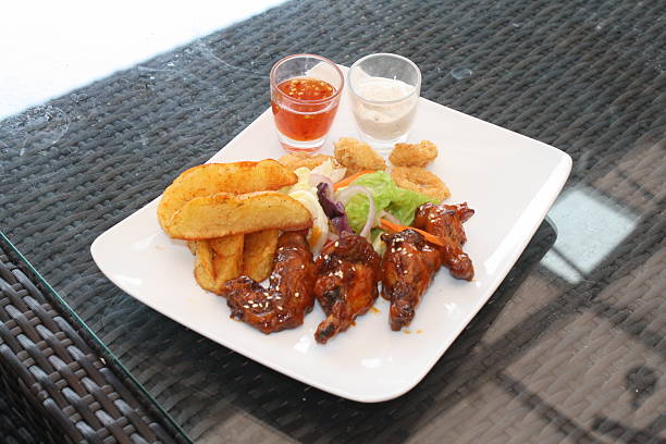 Appetizing plate featuring glazed chicken wings, crispy potato wedges, fresh salad, fried calamari, and two dipping sauces on a glass table