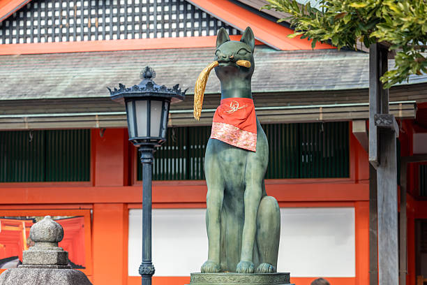 Kyoto, Japan - December 06, 2025: A majestic Inari fox statue, adorned with a red bib and holding a golden scroll, stands guarding Fushimi Inari-taisha shrine, a symbol of Japanese culture and