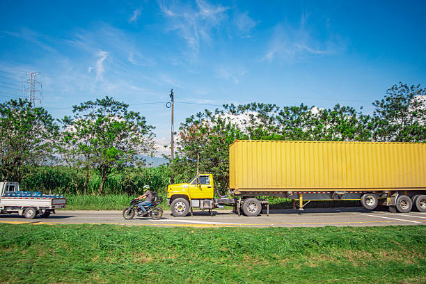 A vibrant scene of a yellow truck and motorcycle on a scenic road in Valle del Cauca, Colombia, surrounded by lush greenery and blue skies.
