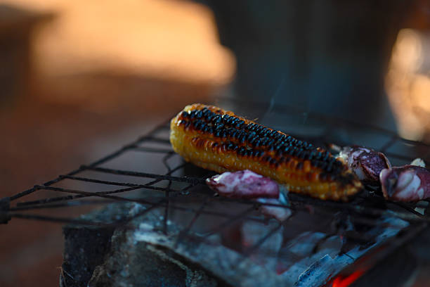A close-up shot of a fresh yellow corn cob being grilled on a traditional charcoal clay stove. The corn is perfectly roasted with some charred spots, emitting soft smoke, creating a delicious and authentic street food atmosphere.