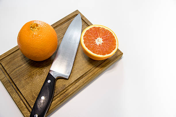 Studio photo of a fresh whole orange and a halved orange placed on a wooden cutting board with a stainless steel kitchen knife. Bright, clean composition with natural orange color and juicy pulp texture, isolated on a white background. Concept of healthy nutrition, fresh fruit, vitamins, food preparation, cooking ingredients, and minimal kitchen still life.