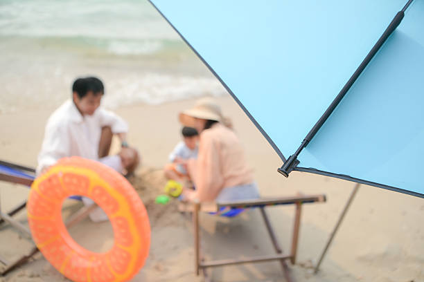 Parents sitting on beach chairs while playing with their young child using sand toys under an umbrella at the beach during daytime