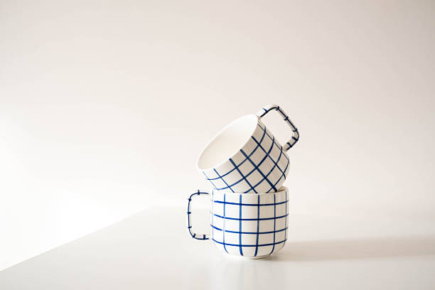 Two white ceramic mugs with a bold blue grid pattern stacked playfully one tilted atop the other. Clean, minimalist background. Perfect for lifestyle or kitchen-themed content. High quality photo
