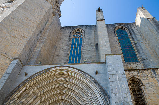 Low angle view of a Gothic cathedral exterior showing tall arched windows, stone walls, and detailed architectural elements under a clear blue sky.