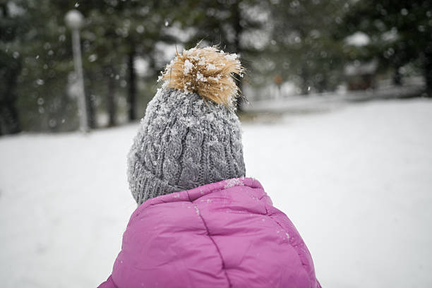 Child from behind enjoying snow falling on hat and jacket in a winter park