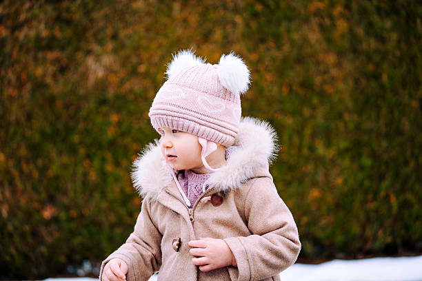 Toddler girl wearing warm winter clothing standing outdoors on a cold day with snow visible in the background.