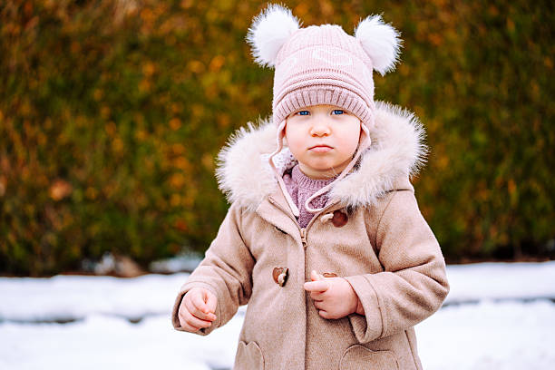 Toddler girl wearing winter clothing looking toward the camera while standing outdoors on a snowy day.