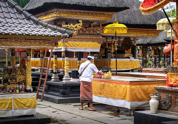 Bali, Indonesia – January 19, 2026: A Balinese man dressed in traditional attire prepares ceremonial offerings at a richly decorated temple shrine. Golden fabrics, carved stone structures, and ceremonial umbrellas frame the scene, illustrating daily Hindu rituals and the spiritual life of Bali.