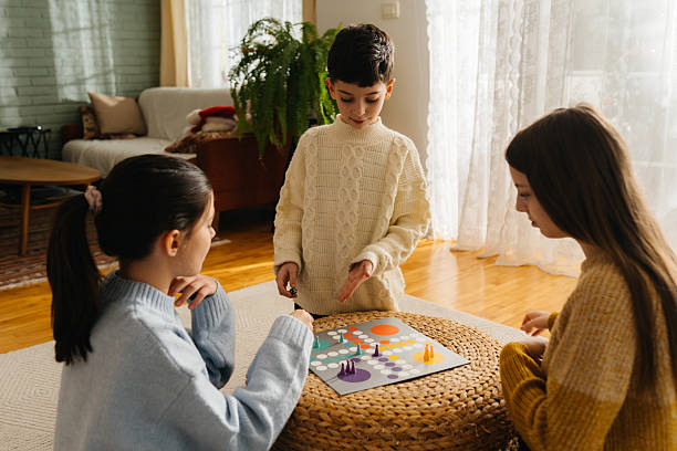 Three children concentrate while playing a board game on a low table indoors. Soft natural light and simple home décor emphasise a calm family activity.