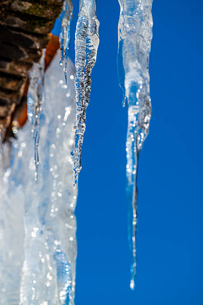 Close-up of clear icicles hanging from a roof edge, frozen water forming long sharp shapes, sunlight reflections on ice, and a deep blue sky background on a cold winter day.
