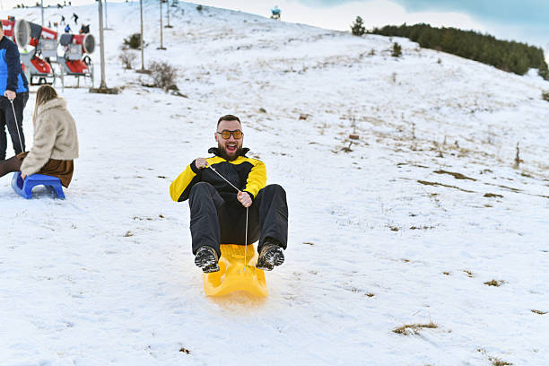 Young man having fun sledding on a snowy slope with a bright yellow sled. Action moment outdoors highlighting winter adventure, leisure, and cold season activities.
