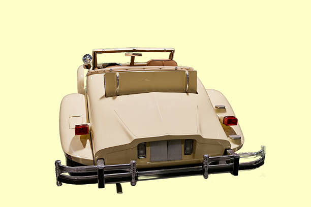 A classic convertible car is parked with its top down. The car has a light color and a smooth design. It is positioned against a plain yellow background.