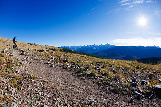 A man rides his electric-powered mountain bike down a singletrack trail in the Rocky Mountains of Alberta, Canada, on a sunny day. He wears cycling clothing and a bicycle helmet.
