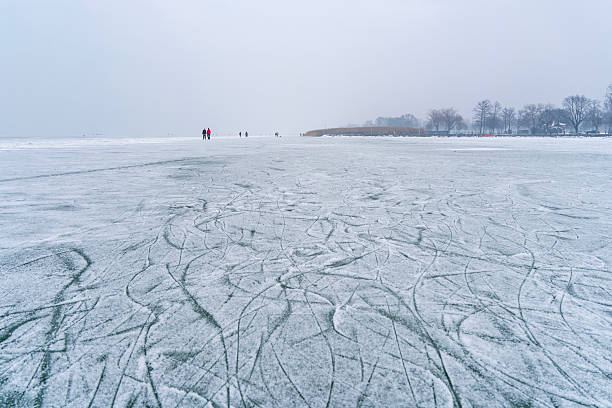 A close-up view of a frozen ice surface covered with numerous intersecting skate marks and scratches. The cool blue tones and textured patterns create an abstract, dynamic winter background associated with ice skating and cold weather.