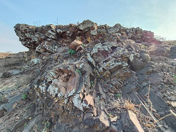 A high-resolution close-up showcasing the rugged, uneven surface of a natural stone specimen. The rock exhibits a complex palette of earthy tones, including deep browns, slate greys, and vibrant reddish-orange oxidation. Its texture is highly granular and fragmented, indicating significant physical and chemical weathering over time.