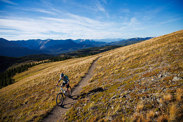 A man rides his full-suspension, electric-powered mountain bike up a singletrack trail in the Rocky Mountains of Alberta, Canada, on a sunny afternoon. He wears cycling clothing, a backpack, and a bicycle helmet.