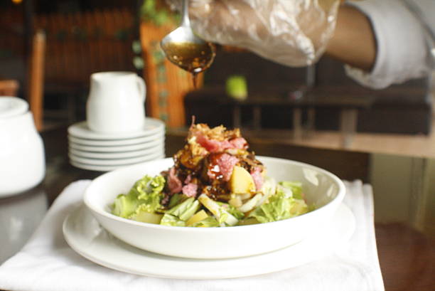 Professional chefs gloved hand carefully pours a rich sauce over a fresh and colorful salad served in a white bowl, ready for dining
