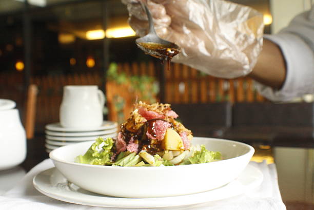Gloved hand meticulously drizzles dark sauce from a spoon onto a fresh gourmet salad with meat and fruit, set in a restaurant dining environment