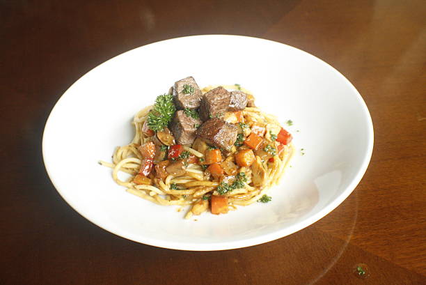 Plated spaghetti topped with savory beef cubes, chopped carrots, mushrooms, and red bell pepper, garnished with fresh parsley on a wooden table