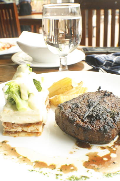 Grilled steak served on a white plate with rich brown sauce, green broccoli, creamy potatoes, and crispy french fries A glass of water is also present
