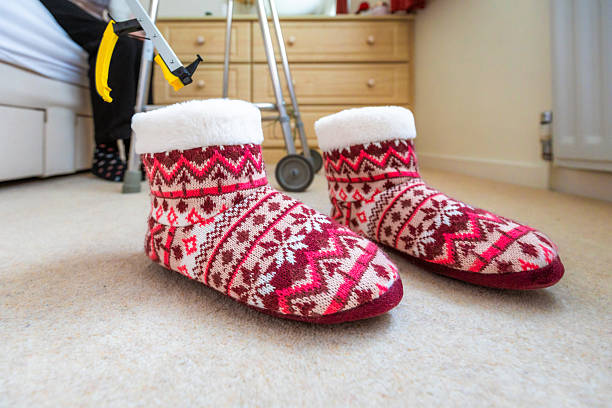 A senior woman in her 70s uses a grabber tool to pick up her slippers from the bedroom floor. The woman uses a mobility walker for support and balance. Focus on the slippers and grabber with the woman defocused.
